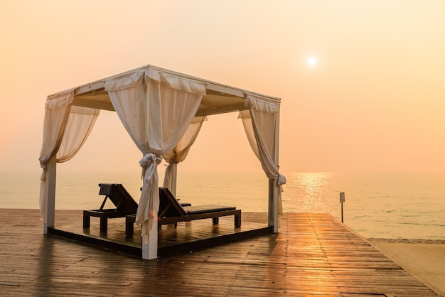 Two lounge chairs under a white cabana on a deck by the sea at sunset.