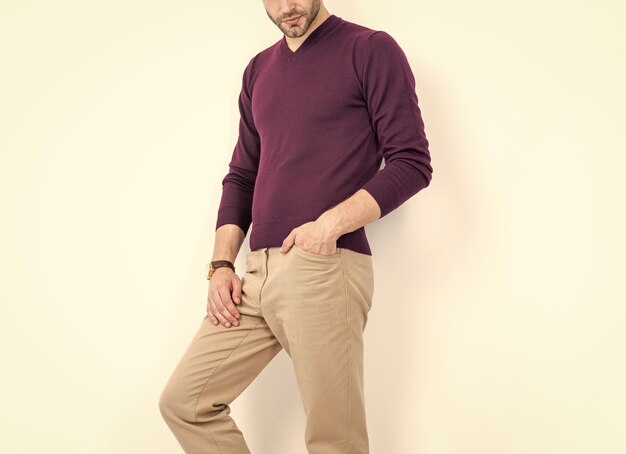 Man wearing a maroon sweater and beige trousers, leaning against a light wall.