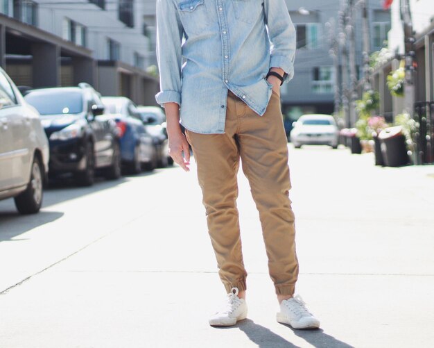 Man in light denim shirt and tan joggers standing on a bright city street, hands in pockets, wearing white sneakers.