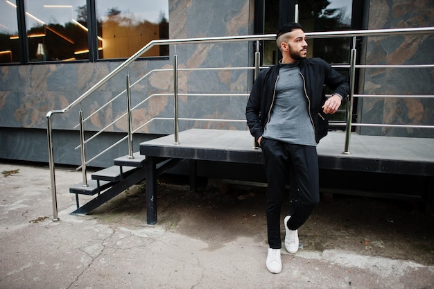 Man in a black jacket, gray t-shirt, and black pants standing outdoors near metal railings, leaning casually with hands in pockets.