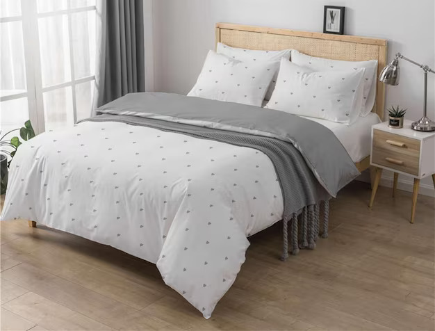 Minimalist bedroom with white patterned duvet, gray throw, and wooden headboard beside a window.